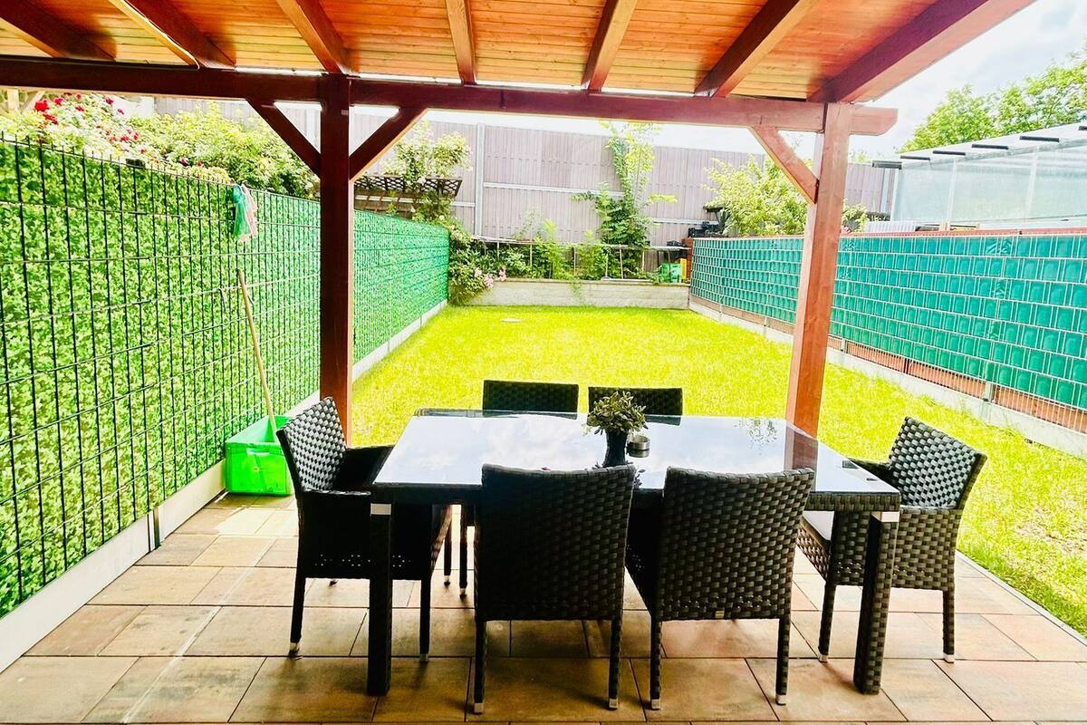 An outdoor patio with a wooden pergola, a table with chairs, and a view of a well-maintained garden and fence.