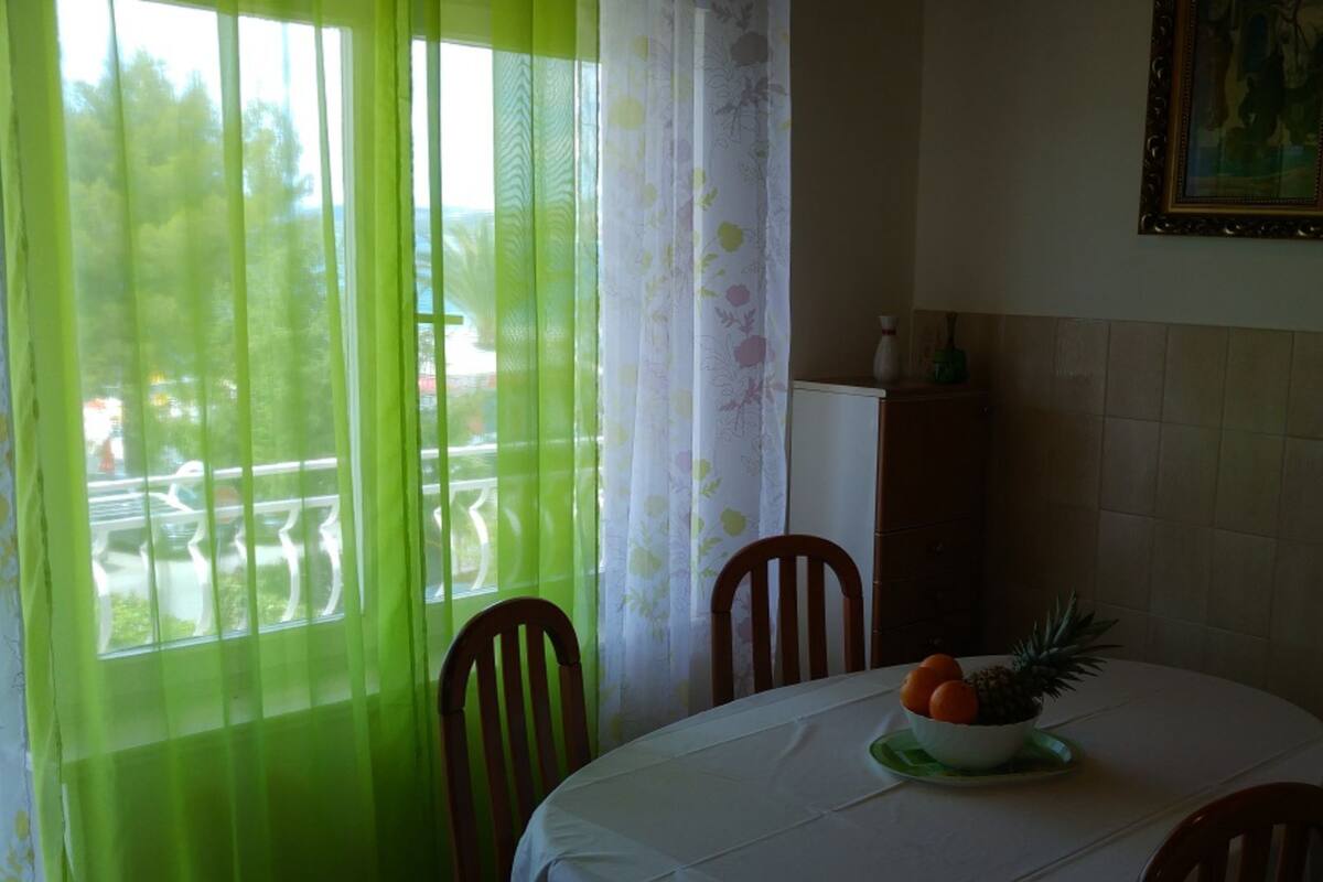 Ein Essbereich mit einem Tisch für zwei Personen, einem Fenster mit grünen Vorhängen und Blick auf eine Straße und Bäume.