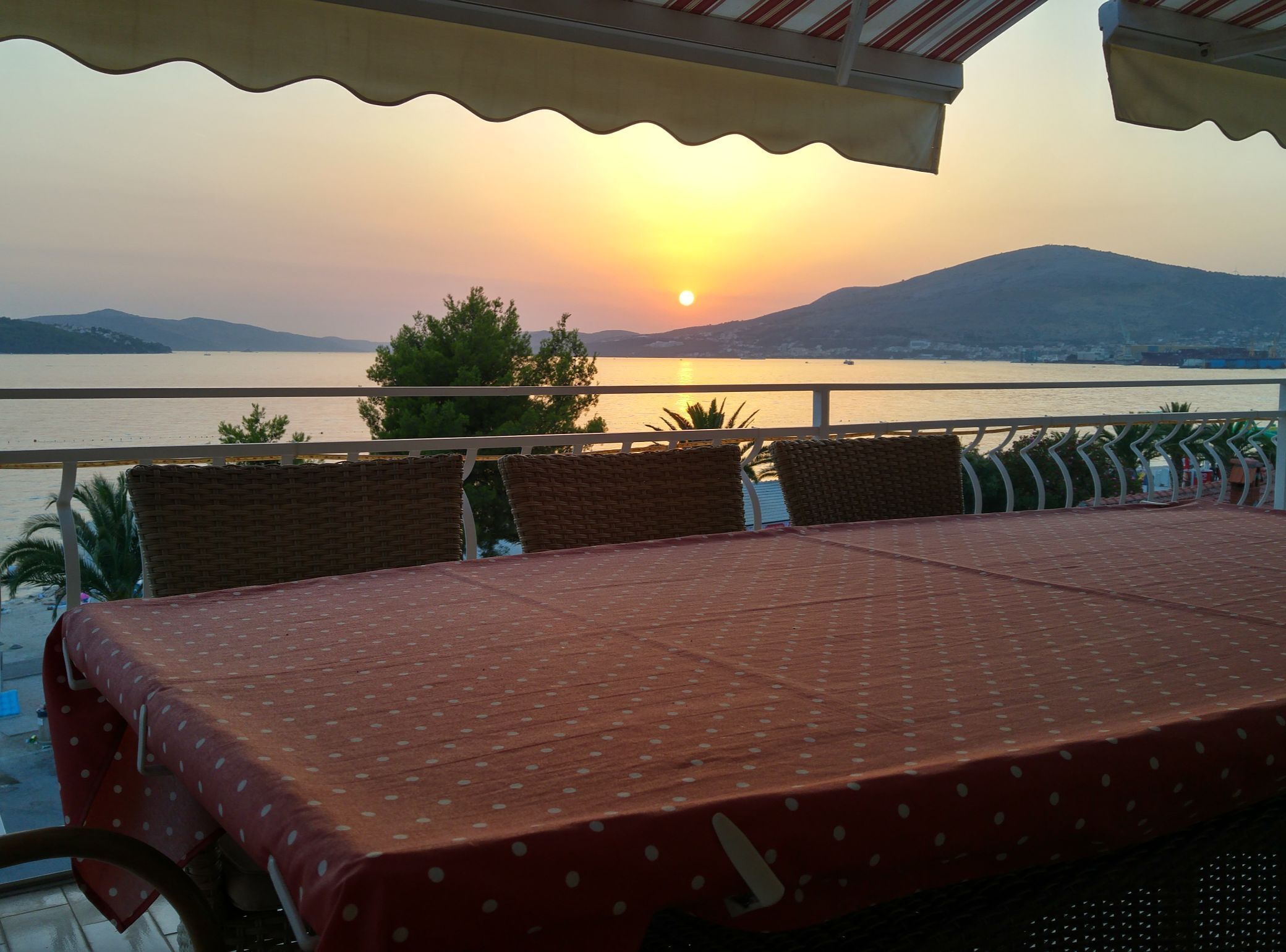 A terrace with a table set for two, overlooking a body of water and distant mountains during sunset.
