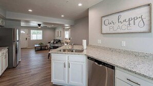 A modern kitchen with granite countertops, stainless steel appliances, and a central island. There is a dining area with a table and chairs, and a living room with a sofa and a coffee table. A framed sign on the wall reads "Our happy place."