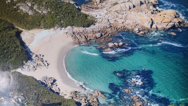 Luchtbeeld van een kustgebied met een zandstrand, rotsformaties en helder turquoise water.