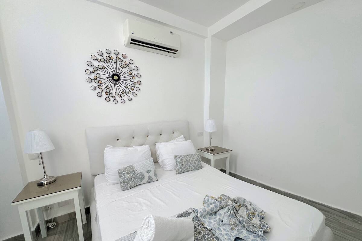 A hotel room with a bed, white walls, an air conditioning unit, a decorative wall piece, a bedside table with a lamp, and a small table with a lamp.