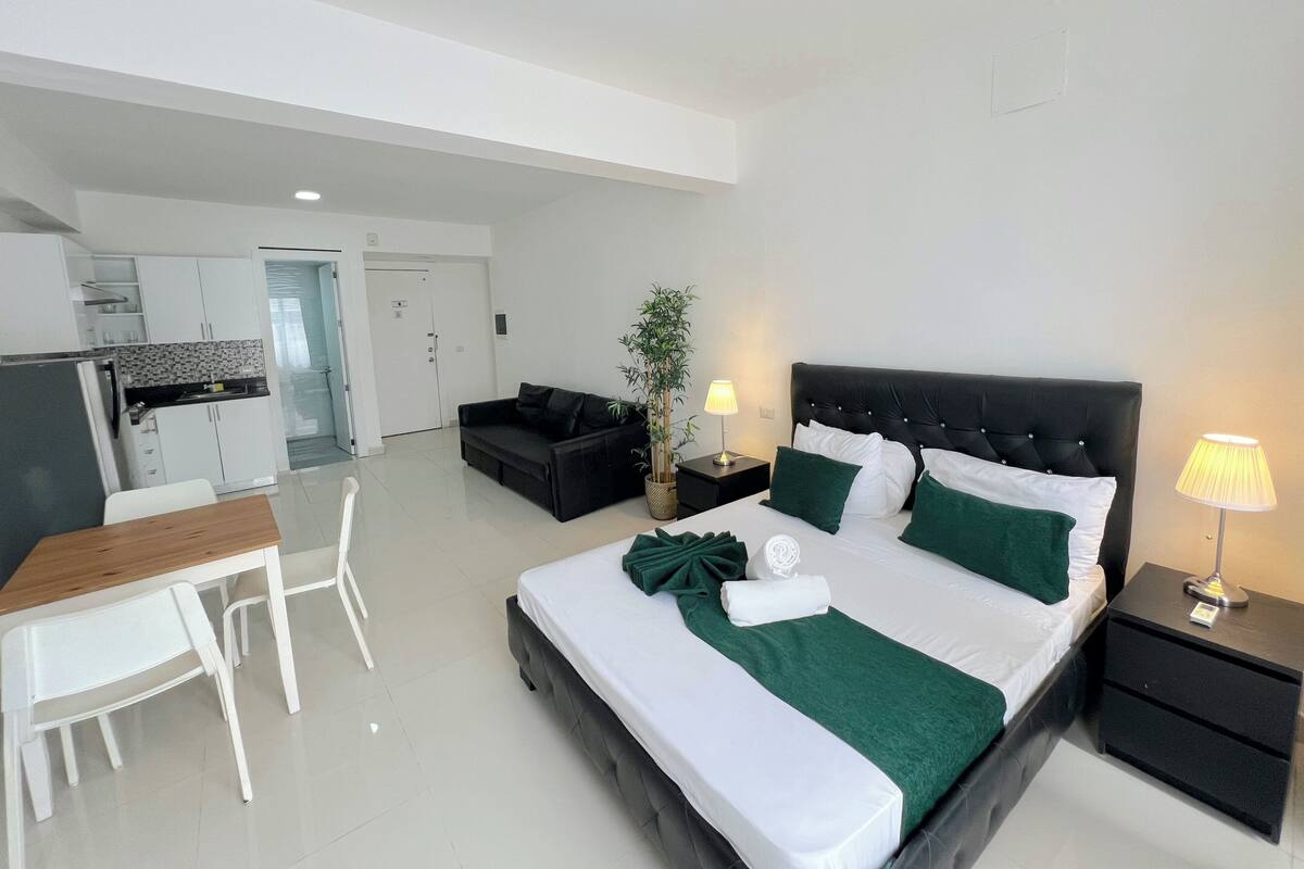 A modern hotel room with a bed, bedside tables, a desk, a chair, and a kitchenette.