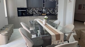 Una zona de menjador moderna amb una taula de vidre, cadires amb coixins i una cuina contemporània al fons.