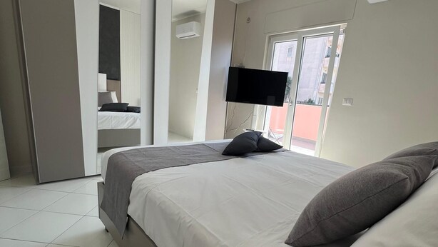 Un dormitori modern amb un llit gran, una televisió de pantalla plana penjada a la paret i una porta corredissa que dona a un balcó.