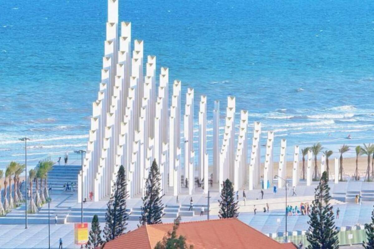 Eine Küstenpromenade mit hohen weißen Säulen, Palmen und einem Strand im Hintergrund.