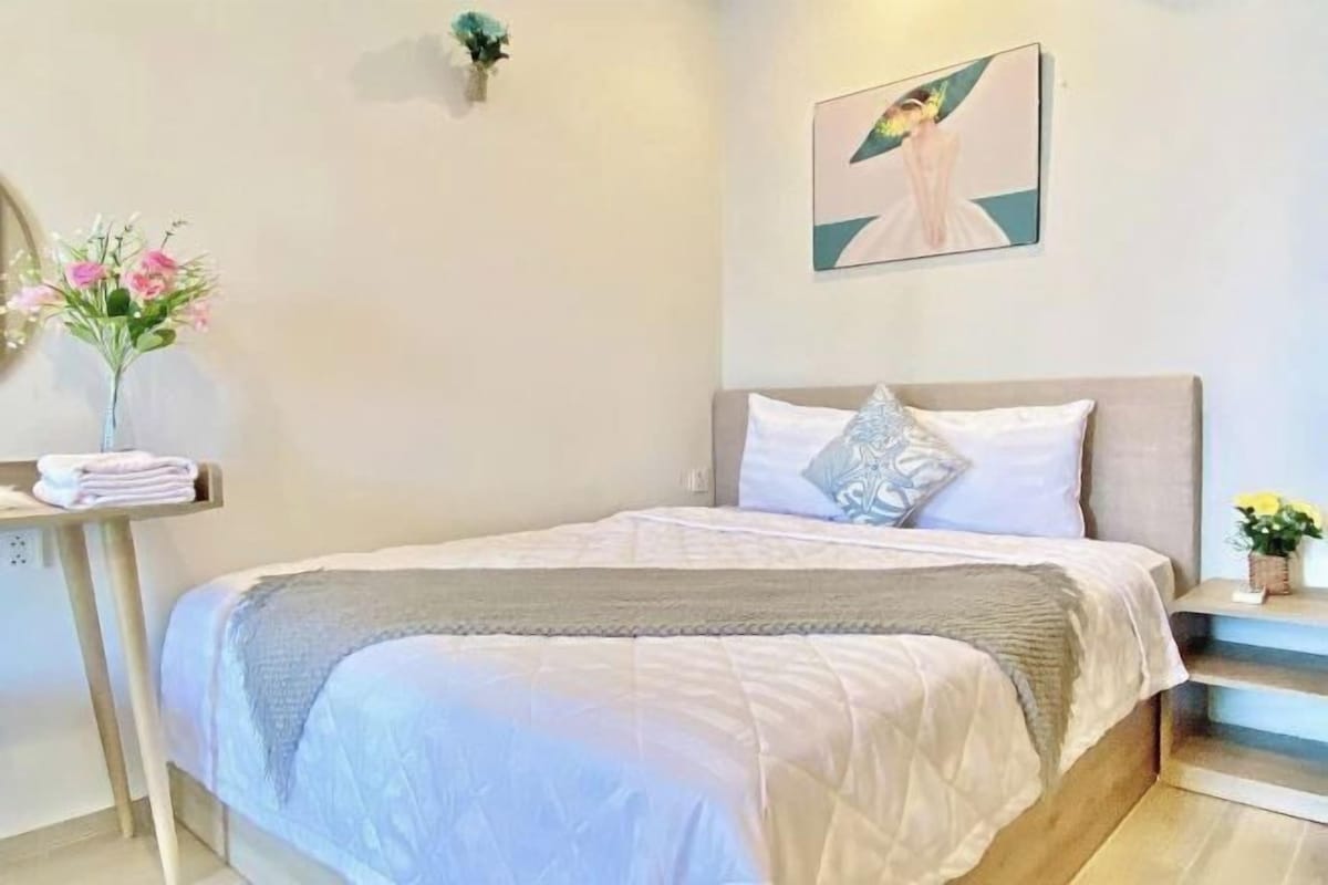Ein Schlafzimmer mit einem Bett, Kissen, einem Nachttisch, einer Vase mit Blumen und einem Bild an der Wand.