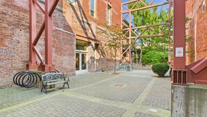 A courtyard with a red metal structure, brick buildings, a bench, and a bicycle rack. - High Ceilings and Modern Loft Living (Victoria Central British Columbia Provincial Government)