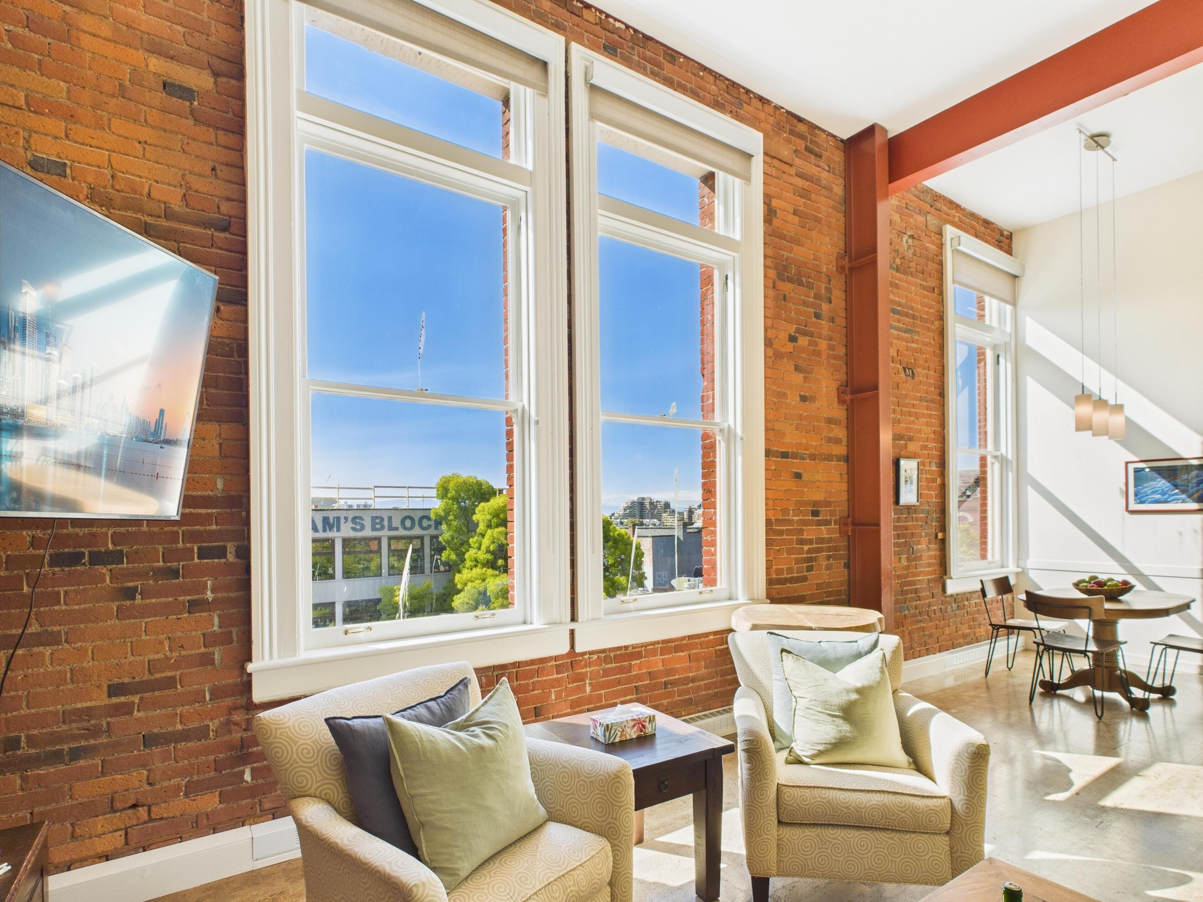 A spacious living area with large windows, a brick wall, and a view of the cityscape.