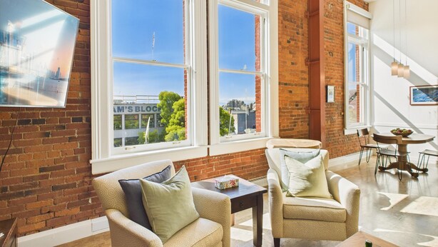 A spacious living area with large windows, a brick wall, and a view of the cityscape. - High Ceilings and Modern Loft Living (Victoria Central British Columbia Provincial Government)