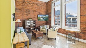 A spacious living room with a brick wall, large windows, a ceiling fan, and a dining area with a table and chairs. - High Ceilings and Modern Loft Living (Victoria Central British Columbia Provincial Government)