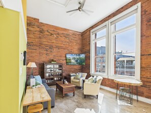 Ein geräumiges Wohnzimmer mit Ziegelwand, großen Fenstern, einem Deckenventilator und einem Essbereich mit Tisch und Stühlen.