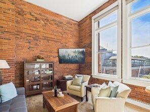 Ein gemütliches Wohnzimmer mit Backsteinwand, großem Fenster, einer Couch, einem Couchtisch und einem Schrank.