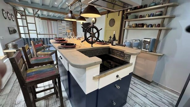 A kitchen with a central island, wooden chairs, and a ship's wheel decoration. - Riva Bella Marine (Ouistreham)