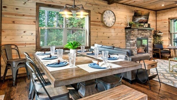 A wooden cabin interior with a dining table set for six, a stone fireplace, and a large window overlooking greenery. - 3 BR • Hot tub • Game Room • Mtn Views • Pools • Golf (Gatlinburg)