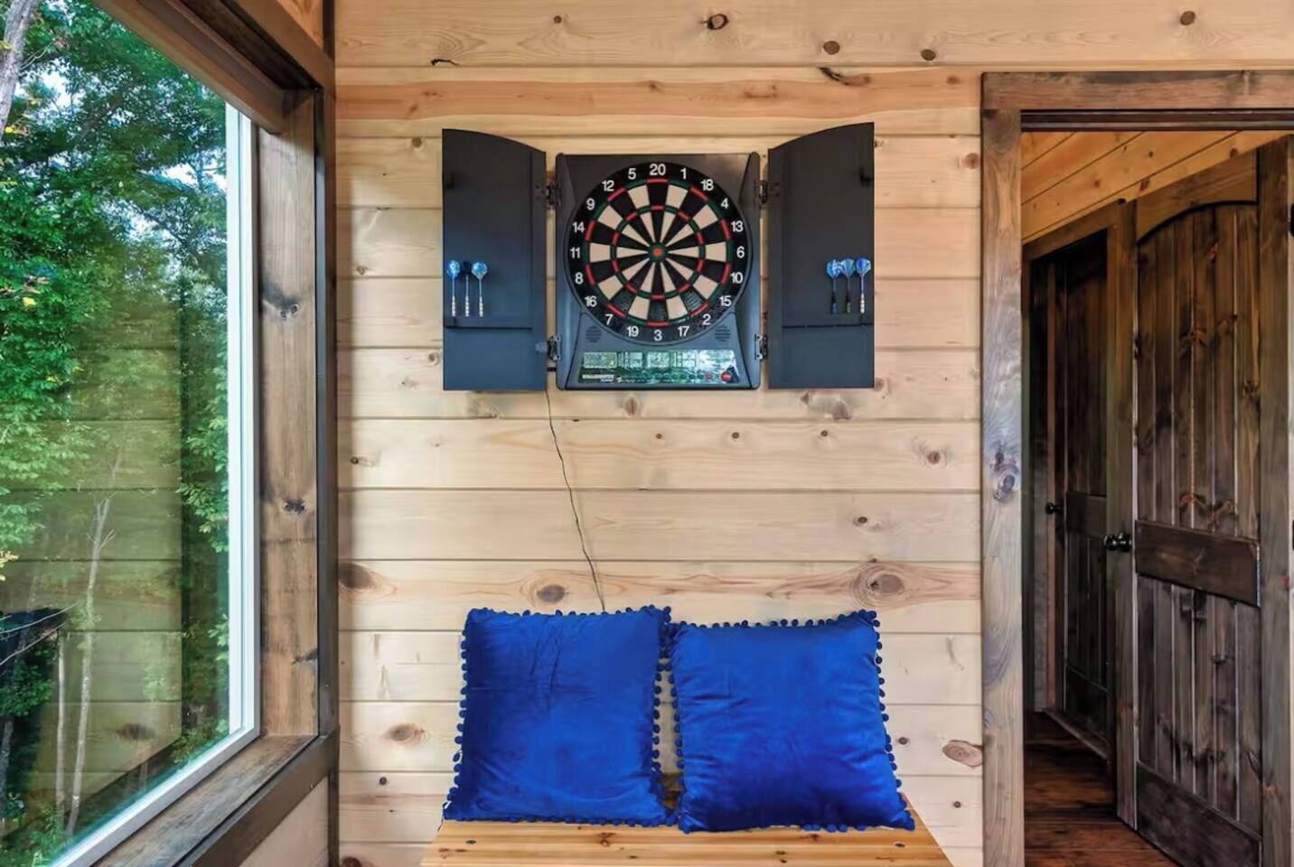 A room with a dartboard mounted on the wall, a bed with blue pillows, and a window providing a view of trees.