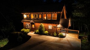 A log cabin with a steeply pitched roof, a wrap-around porch, and a well-lit exterior. - 3 BR • Hot tub • Game Room • Mtn Views • Pools • Golf (Gatlinburg)