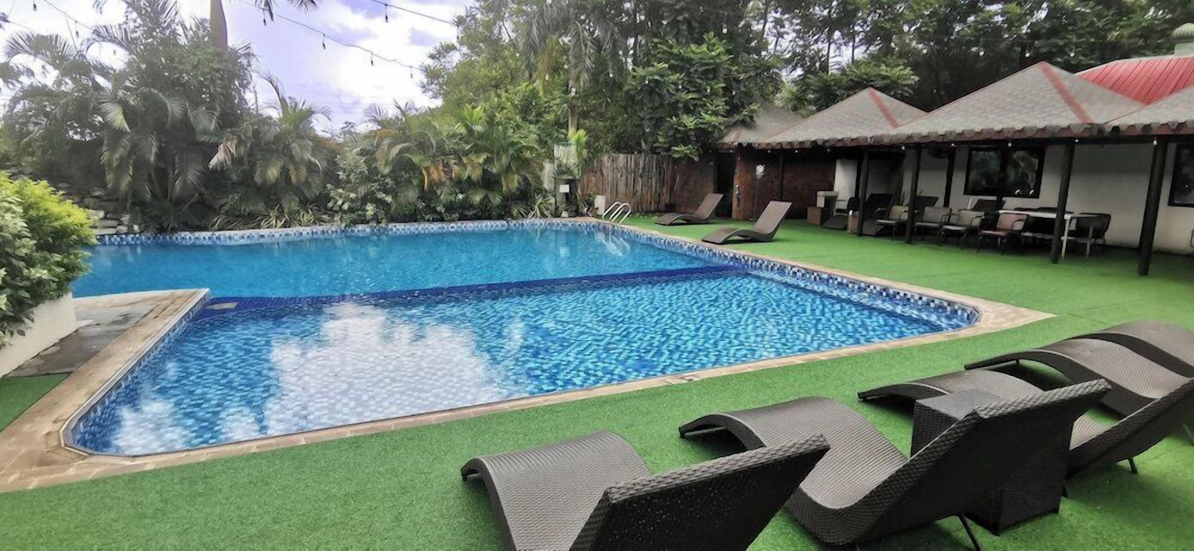 A rectangular swimming pool with a tiled edge, surrounded by green artificial turf. There are lounge chairs and a covered seating area with tables.