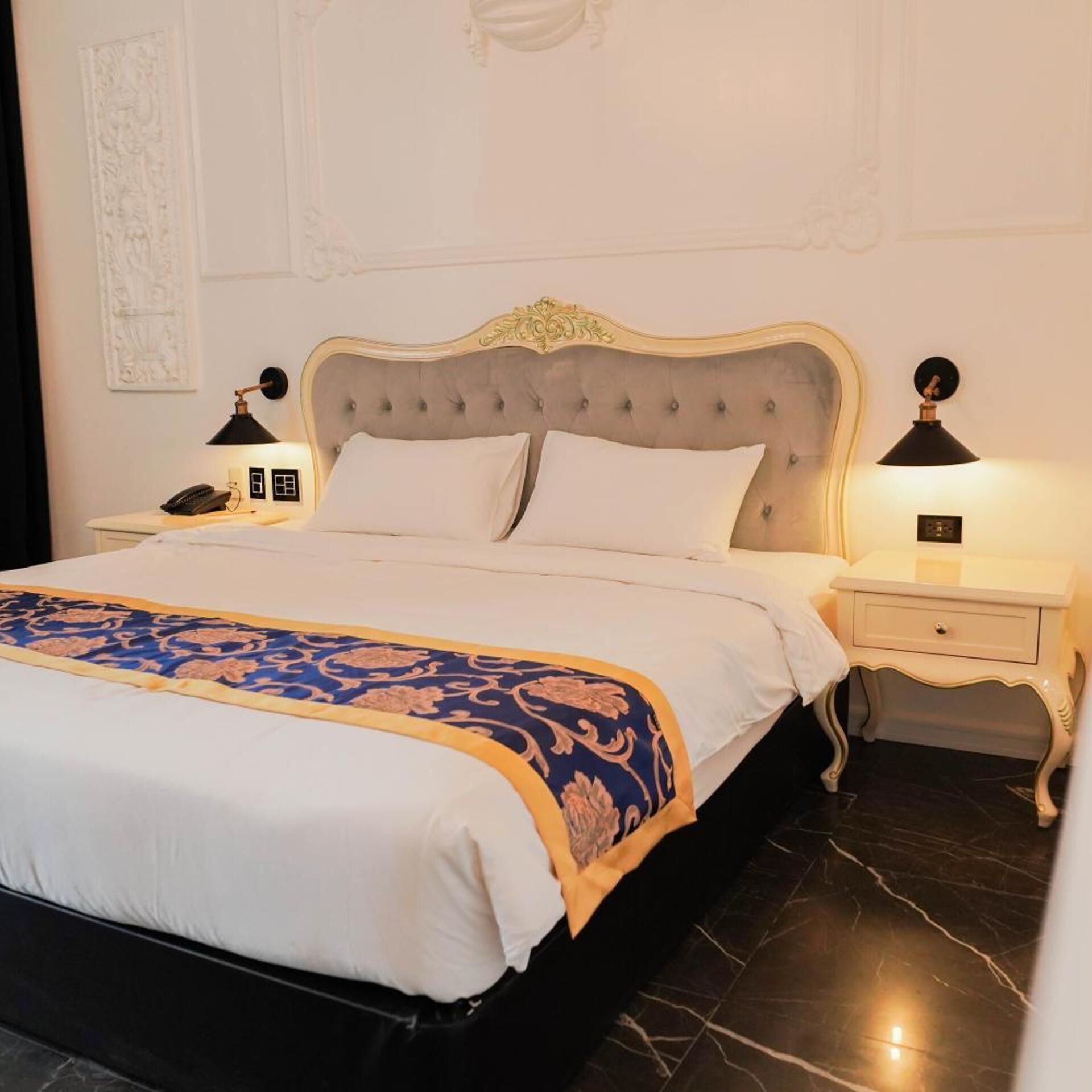 A neatly made bed with a decorative headboard, two bedside tables with lamps, and a marble floor.