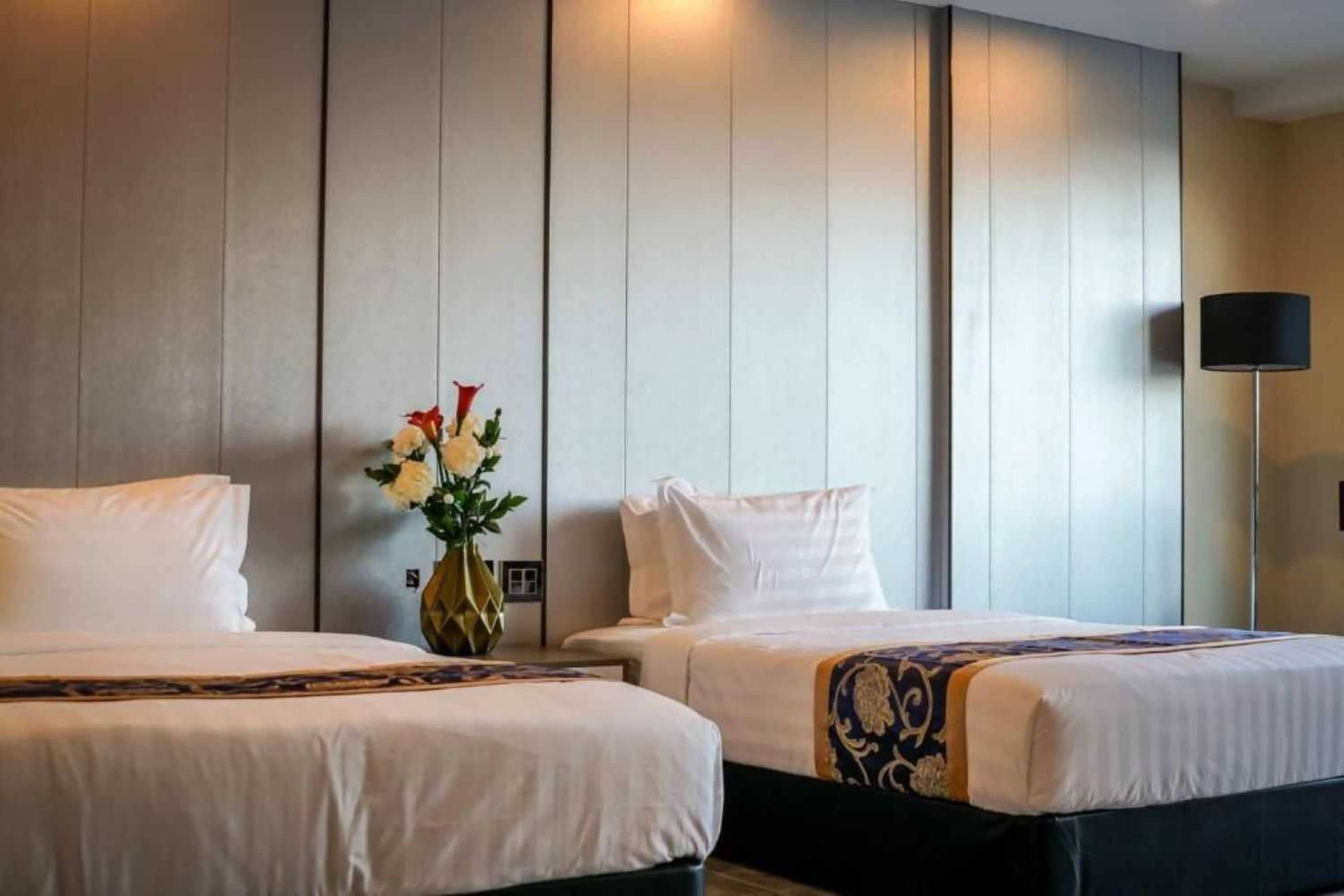 Una camera d'albergo con due letti, un vaso con fiori e una lampada.