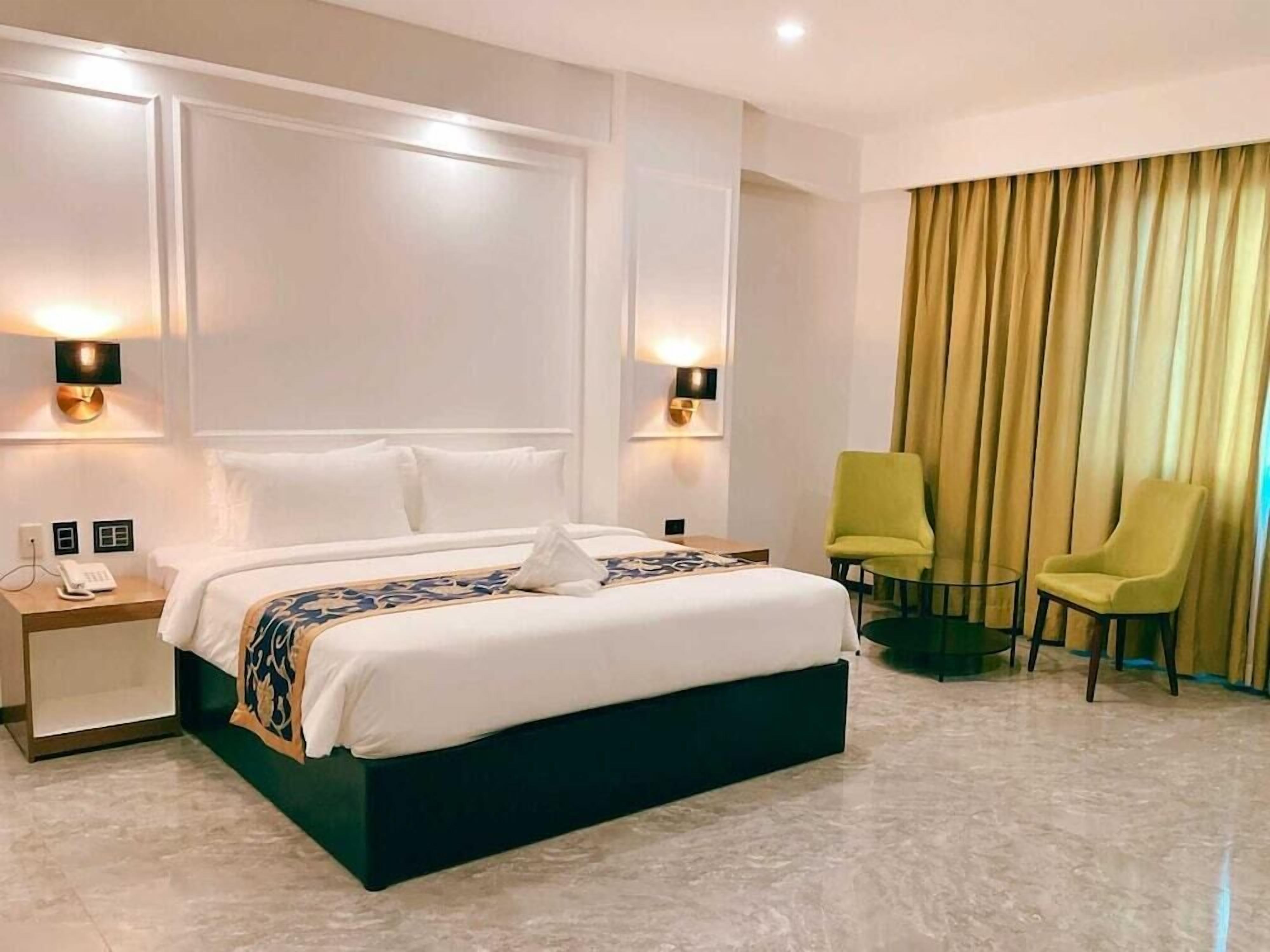 Camera d'hotel con un letto grande, lampade ai comodini, un tavolino, due sedie e una tenda.