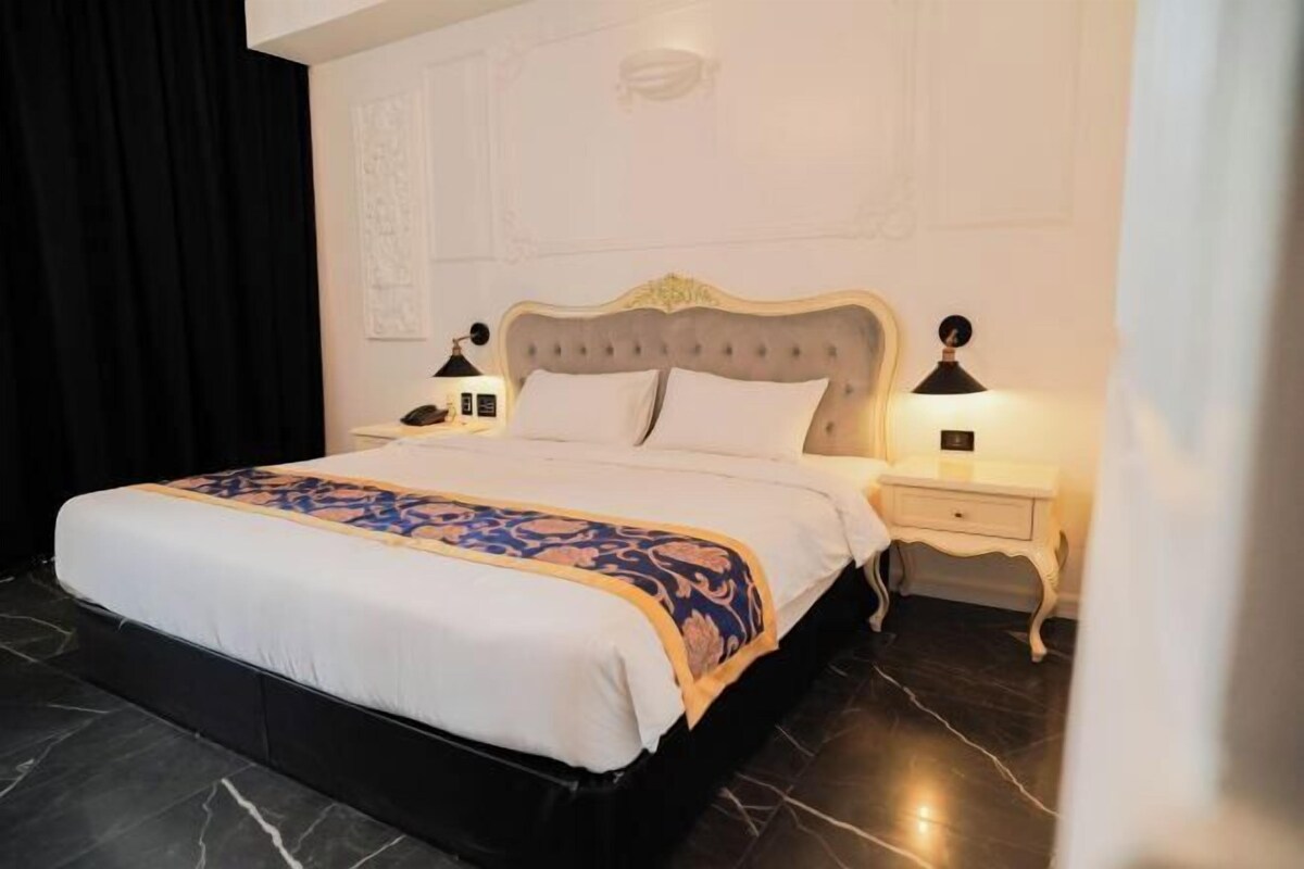 A neatly made bed with a patterned bedspread, a headboard, two bedside tables with lamps, and a dark tiled floor.