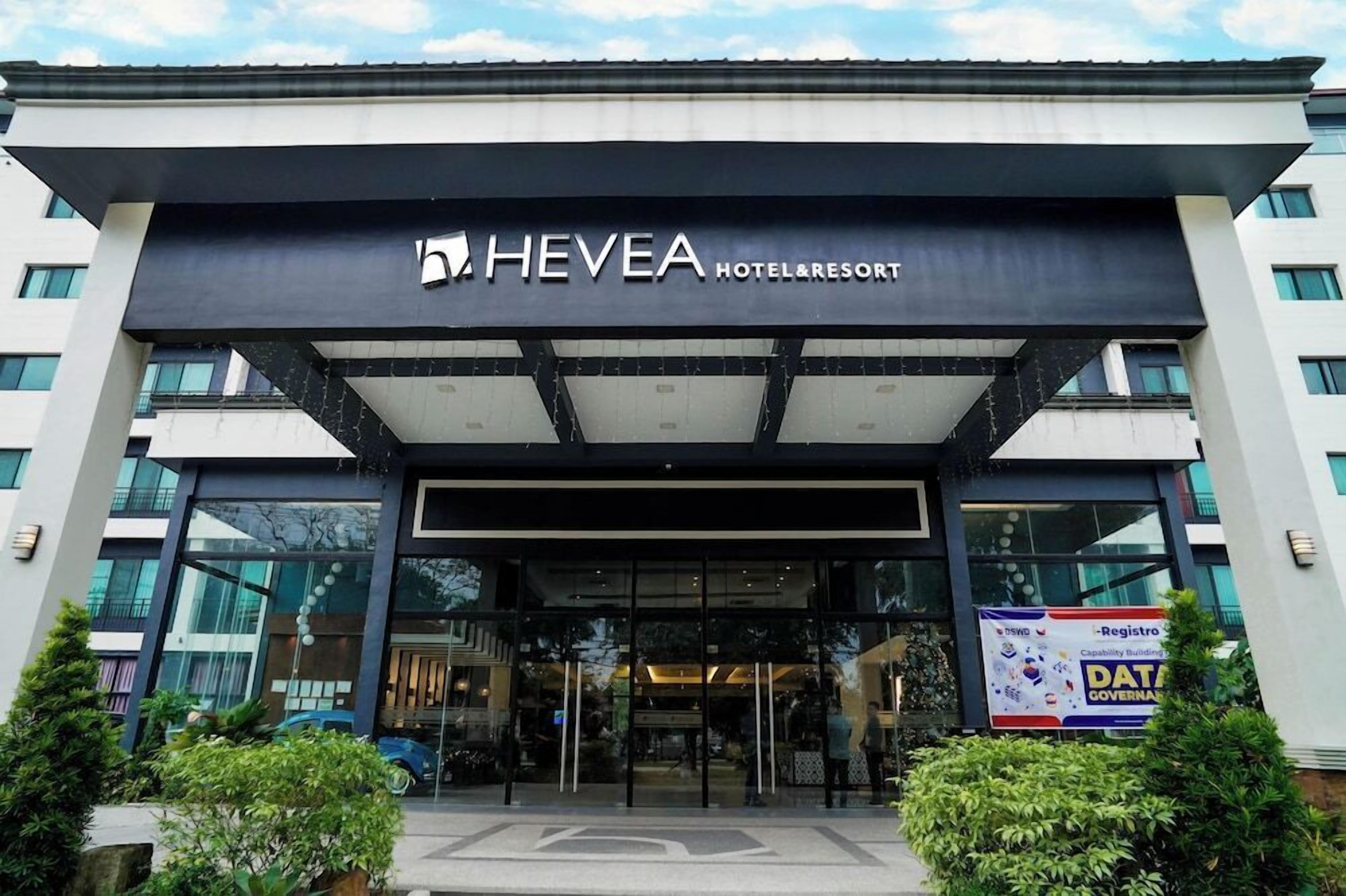 A modern hotel entrance with a glass facade, a prominent sign reading 'HEVEA HOTEL & RESORT,' and a banner for 'DSND - Registro - Capability Building - DATA GOVERNANCE.'