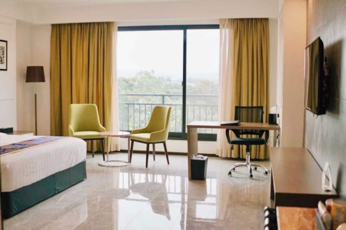 Ein Hotelzimmer mit einem Bett, zwei Stühlen, einem Schreibtisch, einem Fernseher und einem großen Fenster mit Vorhängen.