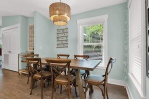 Un coin repas avec une table et des chaises en bois, un luminaire suspendu et une fenêtre avec des stores.