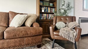 Un sofá de cuero marrón con dos almohadones, un sillón de cuero marrón con un plaid, y una estantería de madera llena de libros.