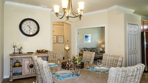 A dining area with a chandelier, wicker chairs, a dining table set for four, and a clock on the wall.