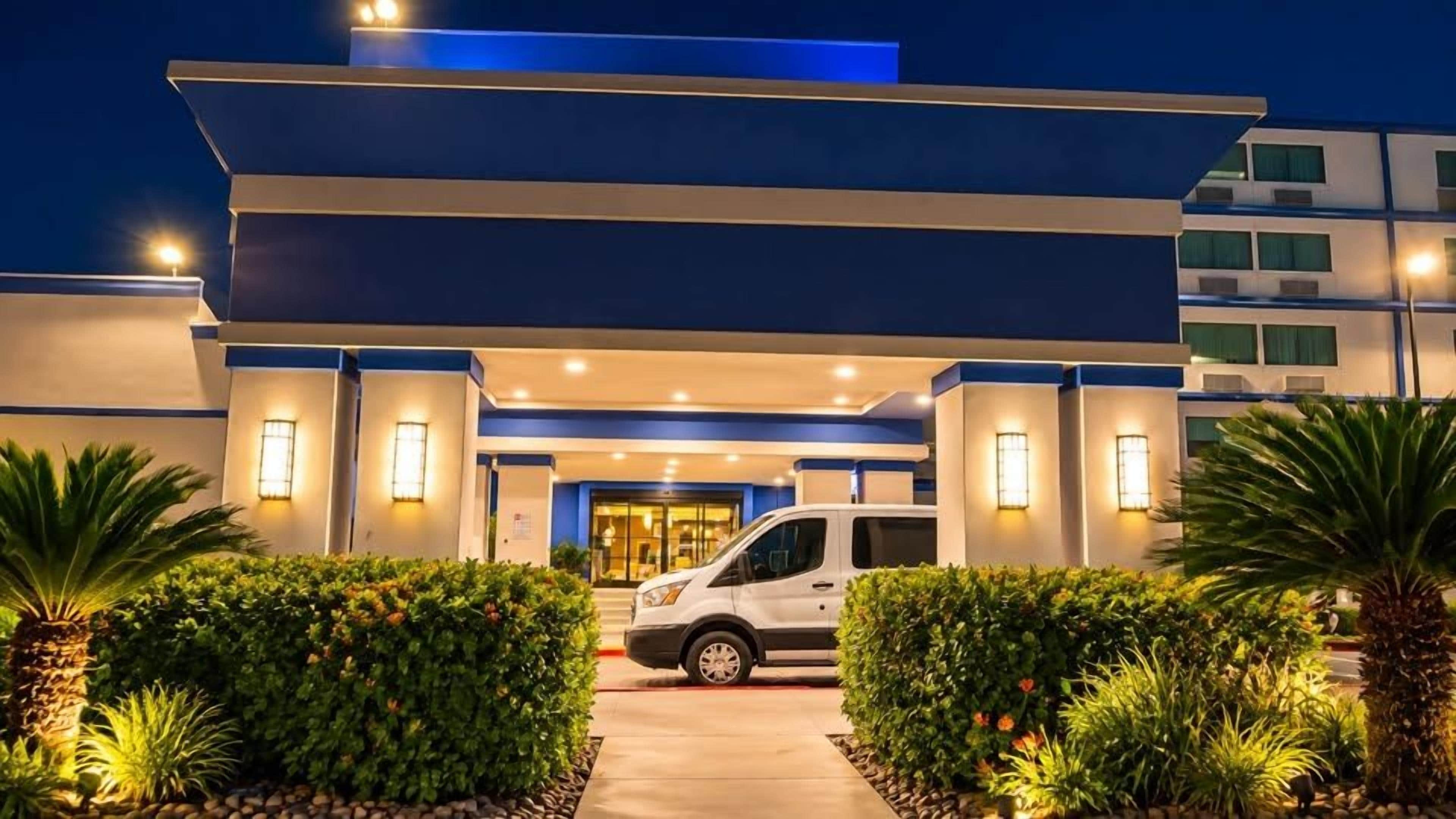 A modern hotel entrance with a white van parked in front, surrounded by well-maintained greenery.