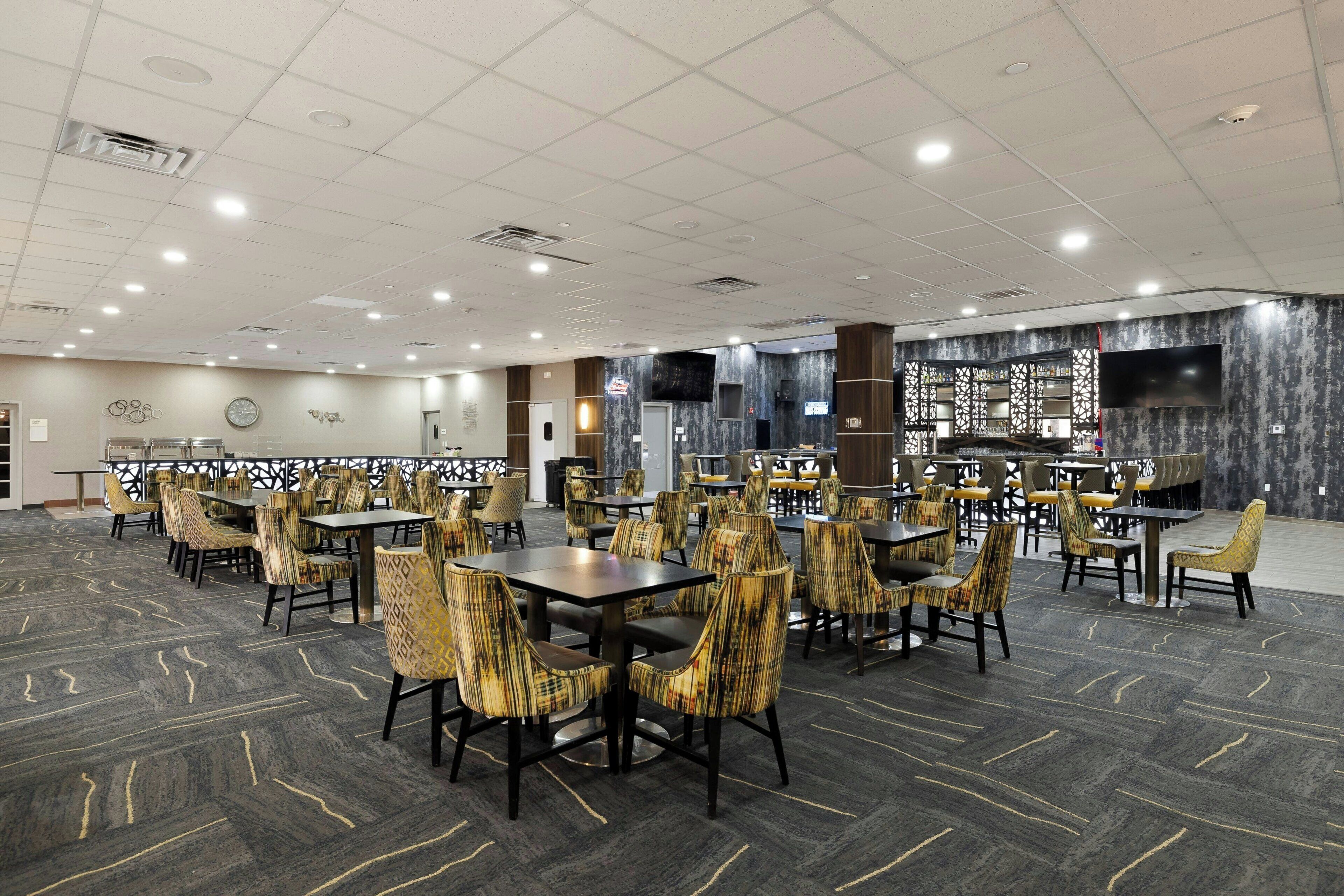 A spacious dining area with multiple tables and chairs, a large flat-screen TV mounted on the wall, and a patterned carpet.