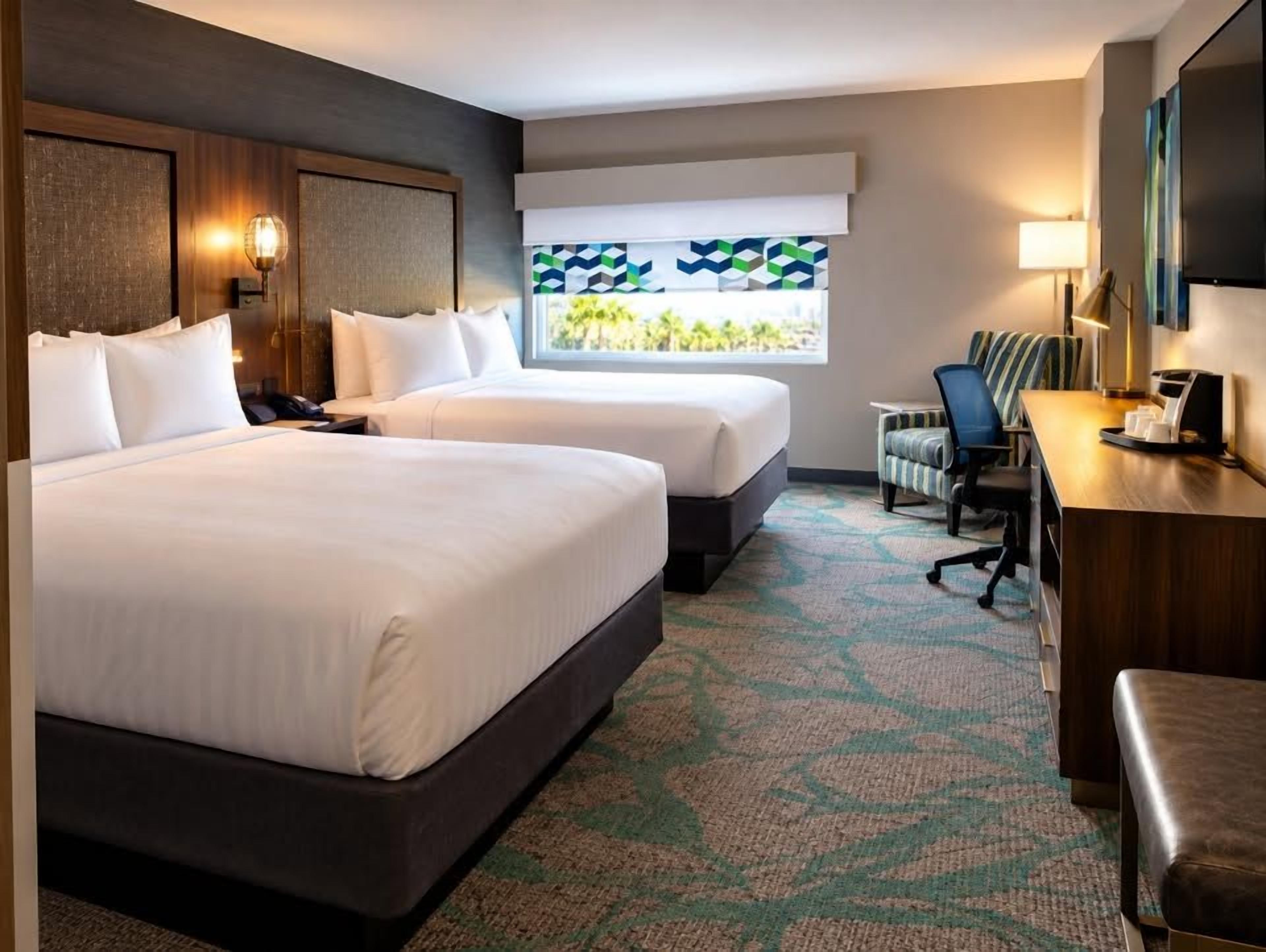 A hotel room with two beds, a desk, a chair, and a TV.