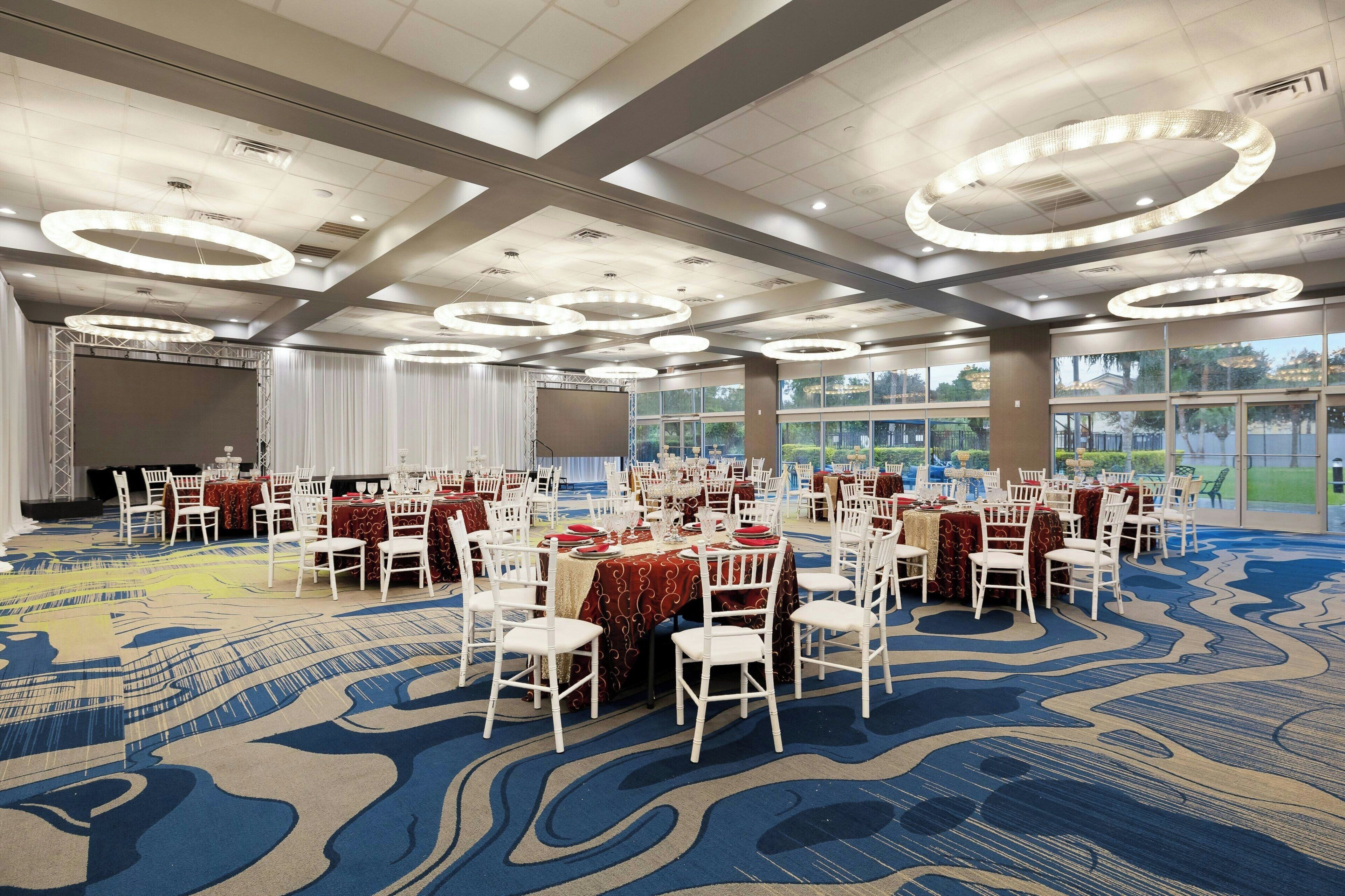 A banquet hall with round tables and white chairs, large windows, and a patterned carpet.