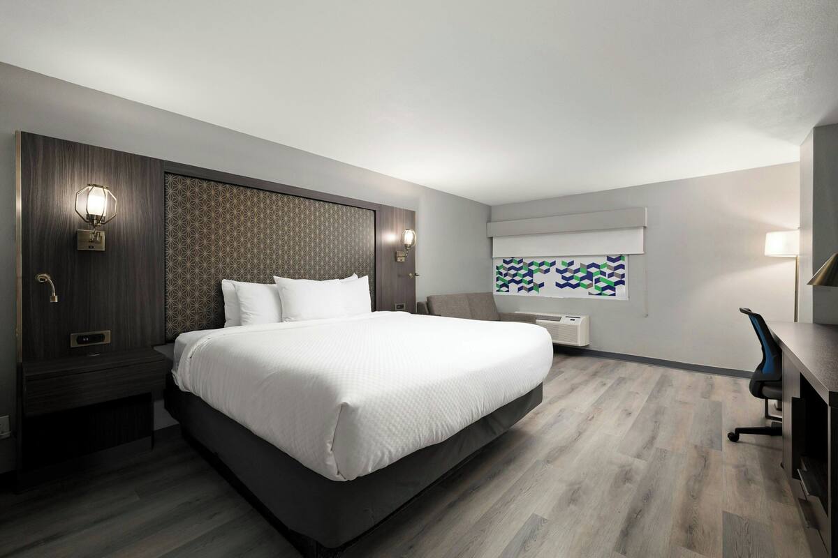 Habitación de hotel moderna con una cama grande, paneles de madera oscura, un escritorio con una silla y una ventana con un diseño geométrico.