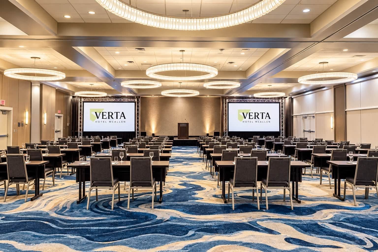 A conference room with rows of tables and chairs, large screens displaying the hotel name, and a blue patterned carpet.