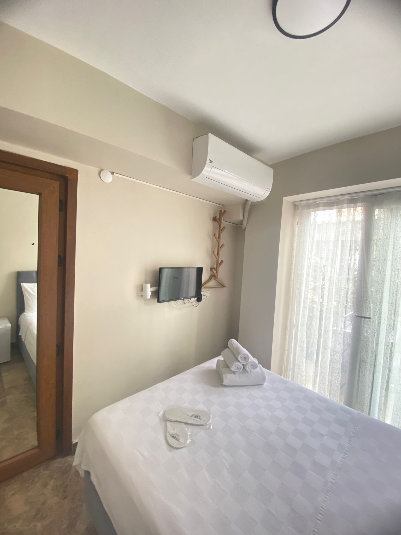 Une chambre d’hôtel comprenant un lit, un téléviseur fixé au mur, un appareil de climatisation, une porte de salle de bain et une fenêtre avec des rideaux.