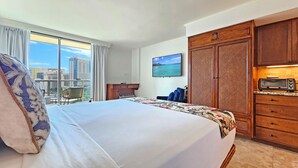 A hotel room with a large bed, a wooden wardrobe, a small kitchenette, and a city view through the window.