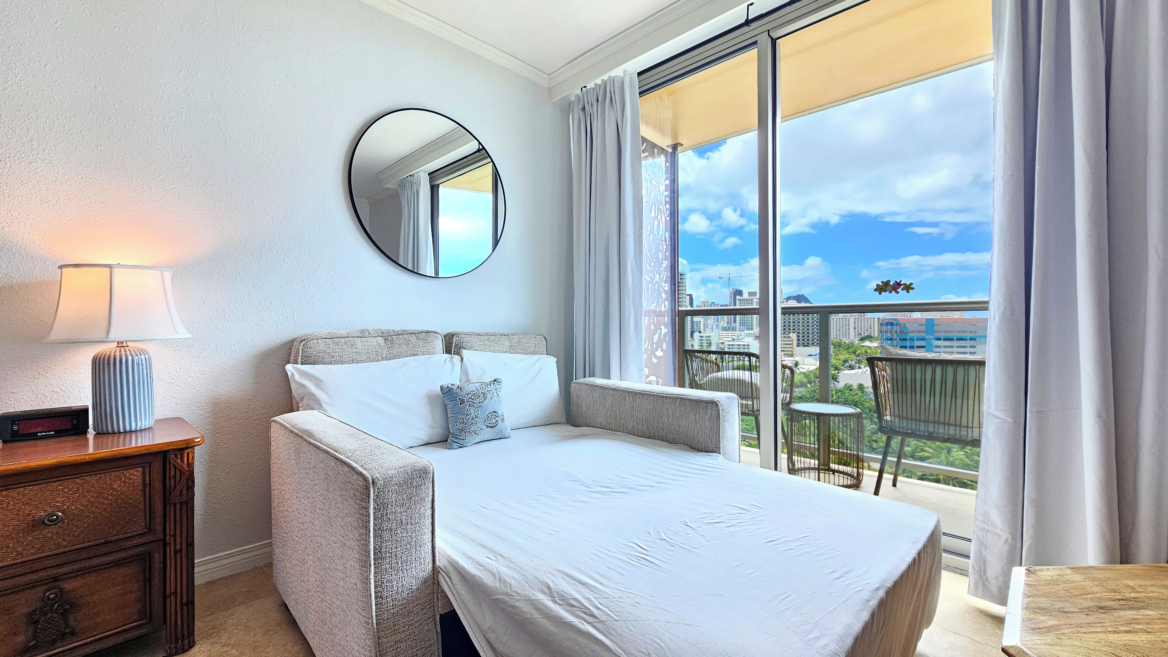 A bedroom with a bed, a bedside table with a lamp, a mirror, and a view of the city through large windows.