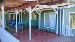 A covered porch with a wooden ceiling and white railings, featuring a seating area with wicker furniture and a grill.