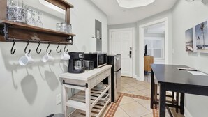A compact kitchen with a coffee maker, microwave, and refrigerator. There is a wooden shelf with glassware and a hook for cups. A dining table is visible with a chair. - Beachside Colony Resort Unit 222A (Tybee Island)