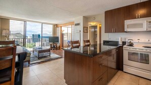 Una cocina moderna con una isla grande, microondas y horno. Hay un comedor con mesa y sillas, y vistas de la ciudad a través de amplias ventanas.