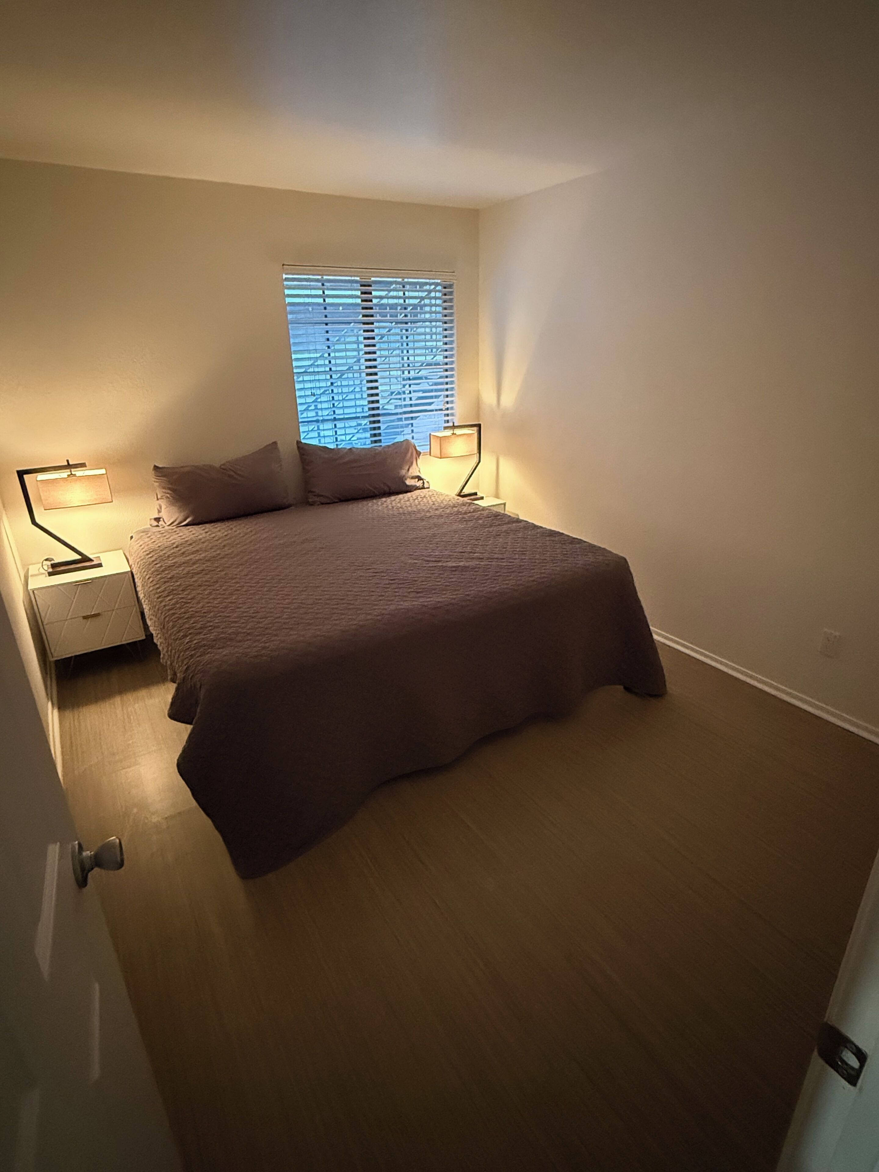 A bedroom with a bed, two bedside tables, a window with blinds, and a door.