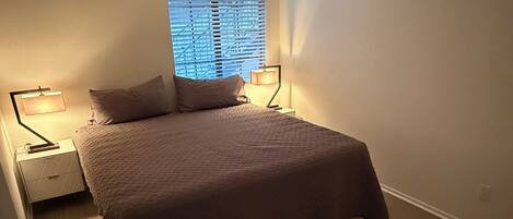A bedroom with a bed, two bedside tables, a window with blinds, and a door.
