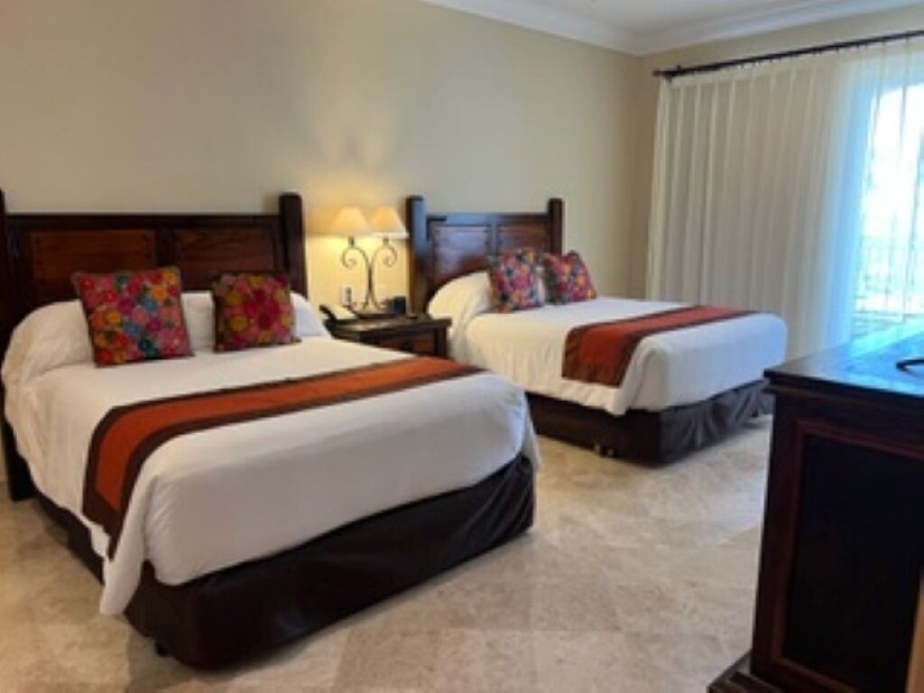 A hotel room with two beds, each with a wooden headboard, white bedding, and colorful pillows. There is a nightstand with a lamp between the beds, and a window with curtains on the right side.