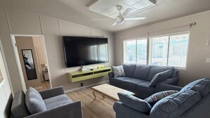 Una sala de estar con una televisión de pantalla plana, dos sofás grises, una mesa de centro y un ventilador de techo.