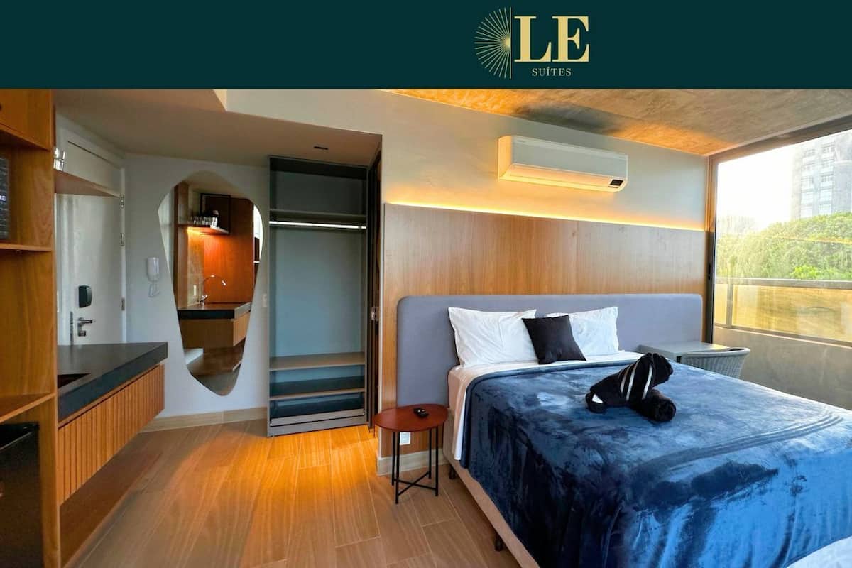 A modern hotel room with a large bed, a small table, a built-in wardrobe, and a window with a city view.