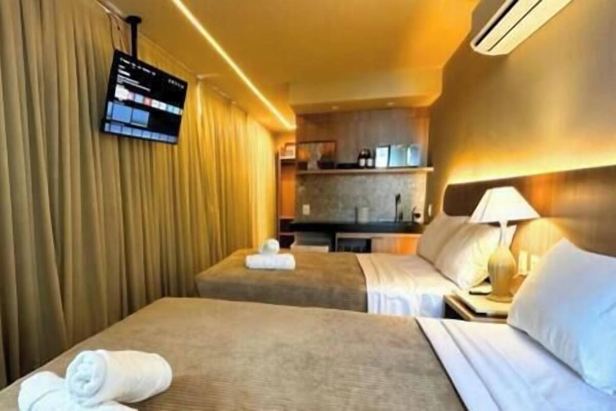 A hotel room with two beds, a TV mounted on the wall, and a kitchenette in the background.