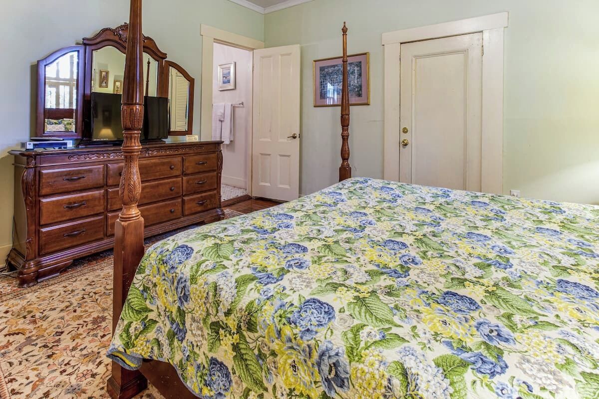A bedroom with a four-poster bed, a wooden dresser, a mirror, and a door.